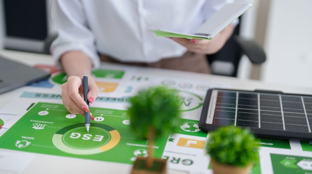 A person at a desk annotating printed sustainability charts with a pen, beside a small plant and a solar panel.